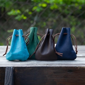 May include: Four small drawstring pouches in teal, green, brown, and navy blue are arranged on a wooden surface. Each pouch has a leather drawstring closure. The background is blurred, with green foliage visible.