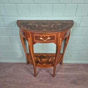 Op de afbeelding: Een halfronde houten bijzettafel met ingewikkelde inlegontwerpen. De tafel heeft een lade met een decoratieve handgreep en een onderste plank. Het hout heeft een warme, roodbruine tint met lichtere accenten, wat een klassieke, elegante stijl laat zien.