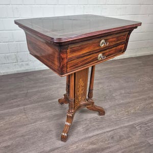 Op de afbeelding: Een kleine, antieke houten bijzettafel met twee lades en een glazen blad. De tafel is gemaakt van donker hout met decoratieve inleg en messing hardware. De tafel is ongeveer 76 cm hoog.