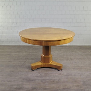 Op de afbeelding: Een ronde, lichtbruine houten tafel met een voetstuk. De tafel heeft een glad, gepolijst oppervlak en een stevige, geometrische voet. De tafel heeft een diameter van ongeveer 91 cm en is ongeveer 76 cm hoog.