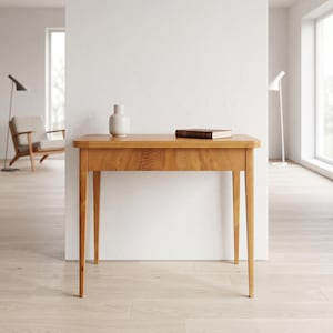 Op de afbeelding: Een lichte houten consoletafel met een rechthoekig blad en vier taps toelopende poten. Een kleine witte vaas en een gesloten boek liggen op de tafel. De tafel is ongeveer 76 cm lang.