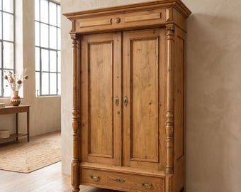 Pode incluir: Um guarda-roupa alto de madeira castanho-claro com duas portas verticais com placas de fechadura de metal simples e uma gaveta horizontal na base com dois puxadores de metal cor de latão. A peça tem colunas decorativas e uma cornija.