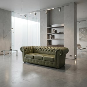Op de afbeelding: Een driezits Chesterfield-bank van olijfgroen leer met knoopdetails en gerolde armen. De bank staat in een moderne, minimalistische woonkamer met betonnen vloeren en muren. De kamer heeft een groot raam en inbouwverlichting.