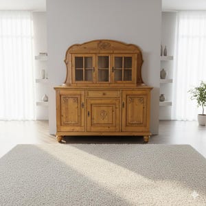 Könnte beinhalten: Ein hellbrauner Holzschrank mit Glastüren und geschnitzten Details. Der Schrank hat eine geschwungene Oberseite und steht auf kurzen, abgerundeten Beinen. Er verfügt über mehrere Schränke, Schubladen und Regale zur Aufbewahrung. Der Schrank steht in einem hellen Raum.