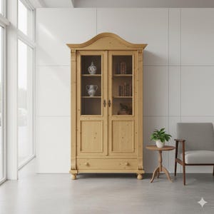 Op de afbeelding: Een lichtgekleurde houten kast met glazen deuren, met decoratieve items en boeken. De kast heeft een gebogen bovenkant en wordt geflankeerd door verticale kolommen. Een kleine ronde tafel en een grijze fauteuil staan in de buurt.