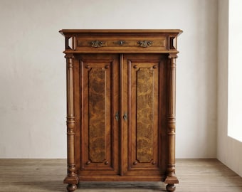 May include: A tall, antique wooden cabinet with two doors and a drawer. The cabinet is made of dark brown wood with a patterned grain. It features ornate hardware and decorative columns on each side. The cabinet stands on a wooden floor.