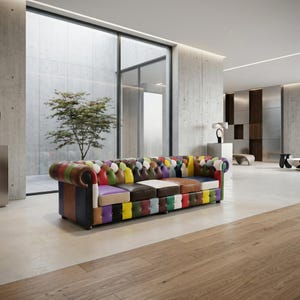 Op de afbeelding: Een grote, kleurrijke Chesterfield-bank met een patchwork-ontwerp. De bank heeft een verscheidenheid aan kleuren, waaronder rood, blauw, groen, geel en paars. De bank staat in een moderne woonkamer met betonnen muren en een groot raam.