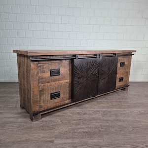 Op de afbeelding: Een rustieke houten mediaconsole met een verweerde afwerking. Het heeft twee lades aan de linkerkant, twee aan de rechterkant en schuifdeuren met een sunburst-ontwerp. De console heeft zwarte metalen hardware en een donkerhouten blad.