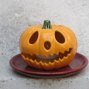 Peut inclure: Une citrouille d'Halloween en céramique orange brillant avec une tige verte, présentant des yeux triangulaires sculptés, un petit nez circulaire et une bouche dentelée, posée sur une assiette en céramique ronde, lisse et bordeaux foncé.