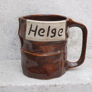 Op de afbeelding: Een bruine keramische mok met een witte band rond de bovenkant. De band heeft de naam "Helge" in bruine letters.