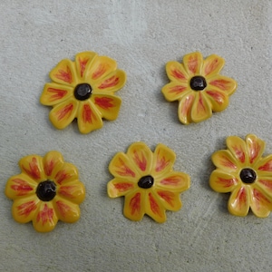 May include: Five ceramic flower decorations in yellow with red petal detailing and dark brown centres. The flowers are displayed on a light grey surface, highlighting their bright colours and intricate design.