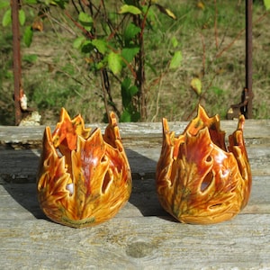 Könnte beinhalten: Zwei Kerzenhalter aus Keramik in Form von herbstlichen Blattclustern in Orange-, Gelb- und Brauntönen. Die Halter haben eine glänzende Oberfläche mit Aussparungen zwischen den Blattformen, was jedem Stück ein strukturiertes, organisches Aussehen verleiht.