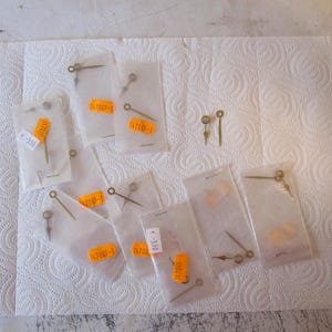 Op de afbeelding: Assortiment van kleine, goudkleurige wijzers voor klokken, verpakt in doorzichtige plastic zakjes met oranje etiketten. De etiketten hebben nummers en een streepje, bijvoorbeeld "17085-1".