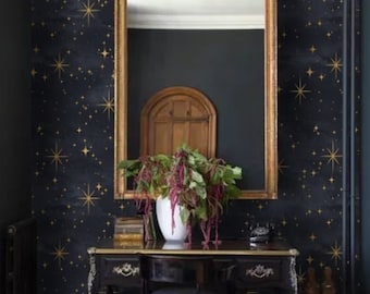 May include: A black desk with a gold trim sits in front of a dark blue wall with a gold star pattern. A white vase with a leafy plant sits on the desk. A mirror with a gold frame hangs on the wall.