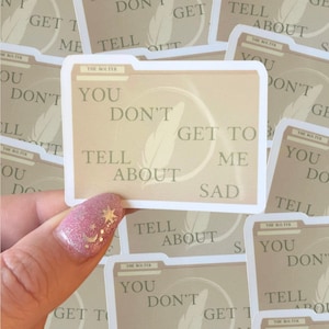 May include: A stack of beige and white stickers with a feather design and the text "You don't get to tell about me sad".