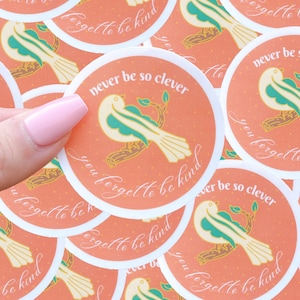 May include: A set of round stickers with a peach background and white border. Each sticker features a white bird with green accents perched on a branch. The text "you forget to be kind" is written in a cursive font around the bird. Some stickers also include the text "never be so clever".