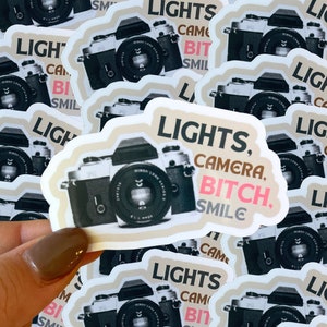 May include: A close-up of a sticker with a vintage camera and the text "LIGHTS, CAMERA, BITCH. SMILE" in pink, black, and brown lettering.
