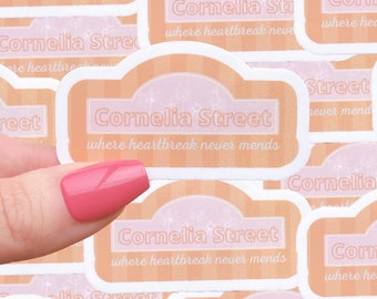 May include: Multiple stickers with the text "Cornelia Street, where heartbreak never mends" in a pink and orange color scheme. The stickers are arranged in a pile, with a hand holding one of the stickers.
