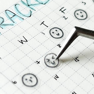 May include: A close-up of a calendar with the word "TRACKER" written at the top. The calendar has a grid pattern and is filled with small, transparent stickers with different facial expressions. A pair of tweezers is holding one of the stickers.