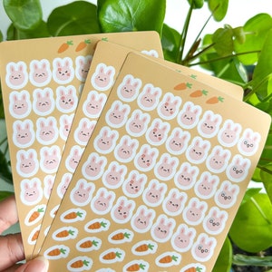 May include: Three sheets of stickers featuring cute bunny faces with different expressions. The stickers are on a light brown background with small orange carrot illustrations. The text on the bottom of the sheet reads "K"