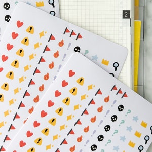 May include: White sticker sheets with colorful icons, including hearts, flags, flames, skulls, and exclamation points. The stickers are arranged in rows and are next to an open planner with grid paper.