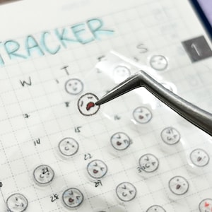 May include: A clear sheet of stickers with various facial expressions. The stickers are arranged in a grid pattern on a white surface. The stickers are designed to be used as a mood tracker.