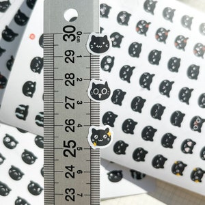 May include: A sheet of black and white cat stickers with different expressions. The stickers are approximately 1 cm in diameter. A ruler is shown for scale.