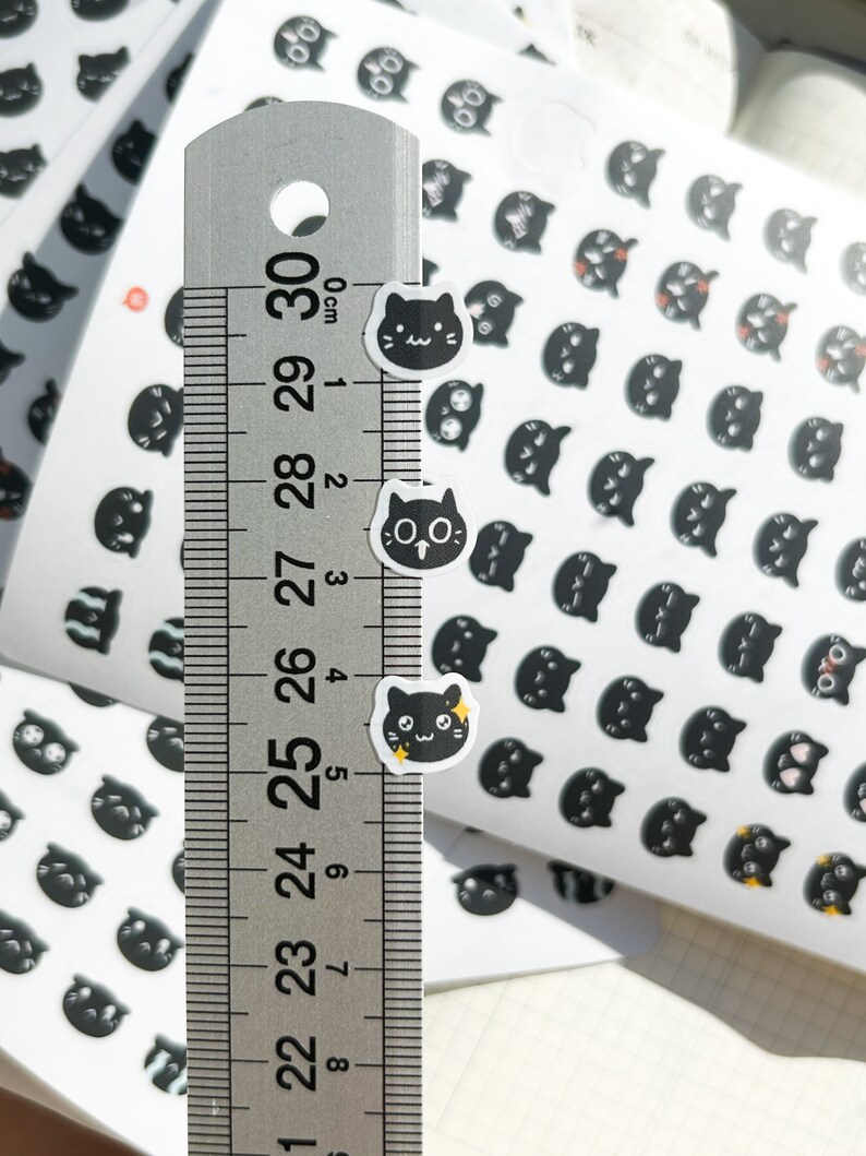 May include: A sheet of black and white cat stickers with different expressions. The stickers are approximately 1 cm in diameter. A ruler is shown for scale.