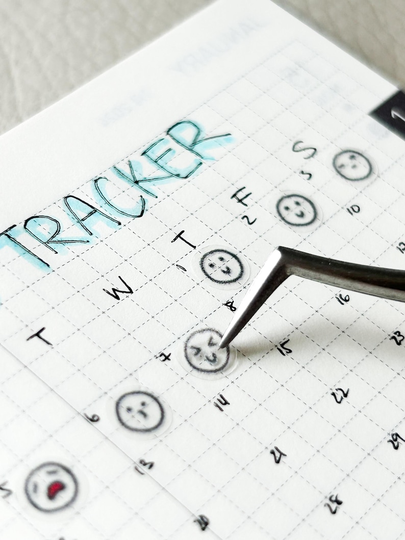 May include: A close-up of a calendar with the word "TRACKER" written at the top. The calendar has a grid pattern and is filled with small, transparent stickers with different facial expressions. A pair of tweezers is holding one of the stickers.