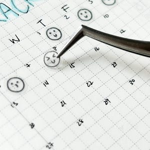 May include: A close-up of a weekly planner with a grid pattern. The planner has a "tracker" heading at the top and numbers 1 through 30 written in the squares. There are small smiley face stickers placed on some of the squares.