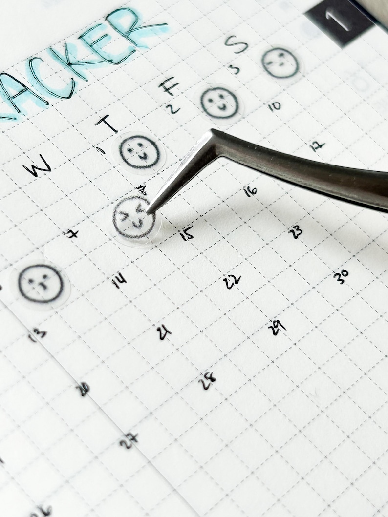 May include: A close-up of a weekly planner with a grid pattern. The planner has a "tracker" heading at the top and numbers 1 through 30 written in the squares. There are small smiley face stickers placed on some of the squares.