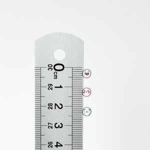 May include: Three small white stickers with different facial expressions. The stickers are placed on a ruler with centimeter markings. The ruler shows measurements from 25 cm to 30 cm.