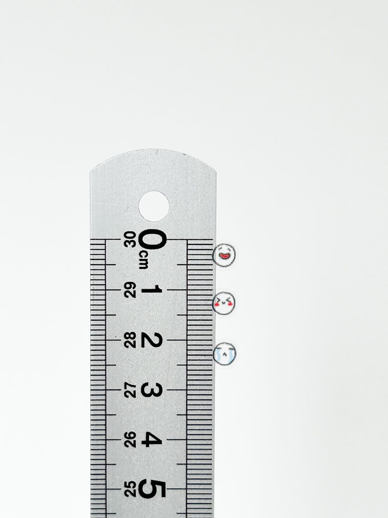 May include: Three small white stickers with different facial expressions. The stickers are placed on a ruler with centimeter markings. The ruler shows measurements from 25 cm to 30 cm.