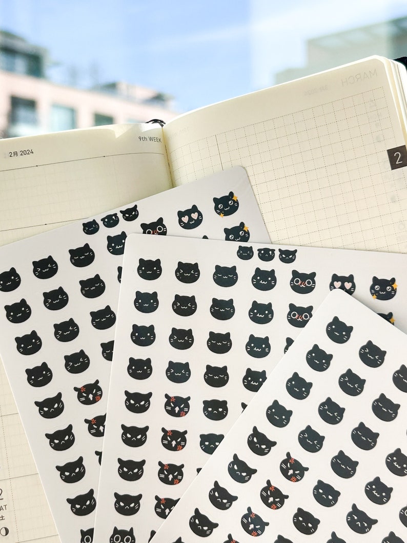 May include: A sheet of black cat stickers with different facial expressions. The stickers are on a white background and are arranged in rows. The stickers are perfect for adding a touch of whimsy to planners, journals, and other paper goods.