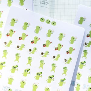 Puede incluir: Una hoja de pegatinas con un personaje de dibujos animados verde en varias poses. El personaje lleva una bolsa de la compra, un maletín, una taza de café y una paleta de hielo. El personaje también lleva un sombrero y gafas de sol. Las pegatinas están impresas en papel blanco y tienen un acabado mate.