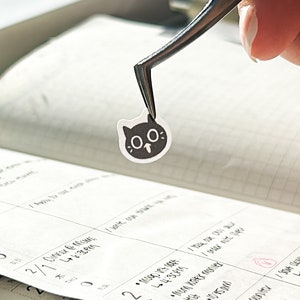May include: A black and white cat sticker with large eyes being held by tweezers over a calendar.