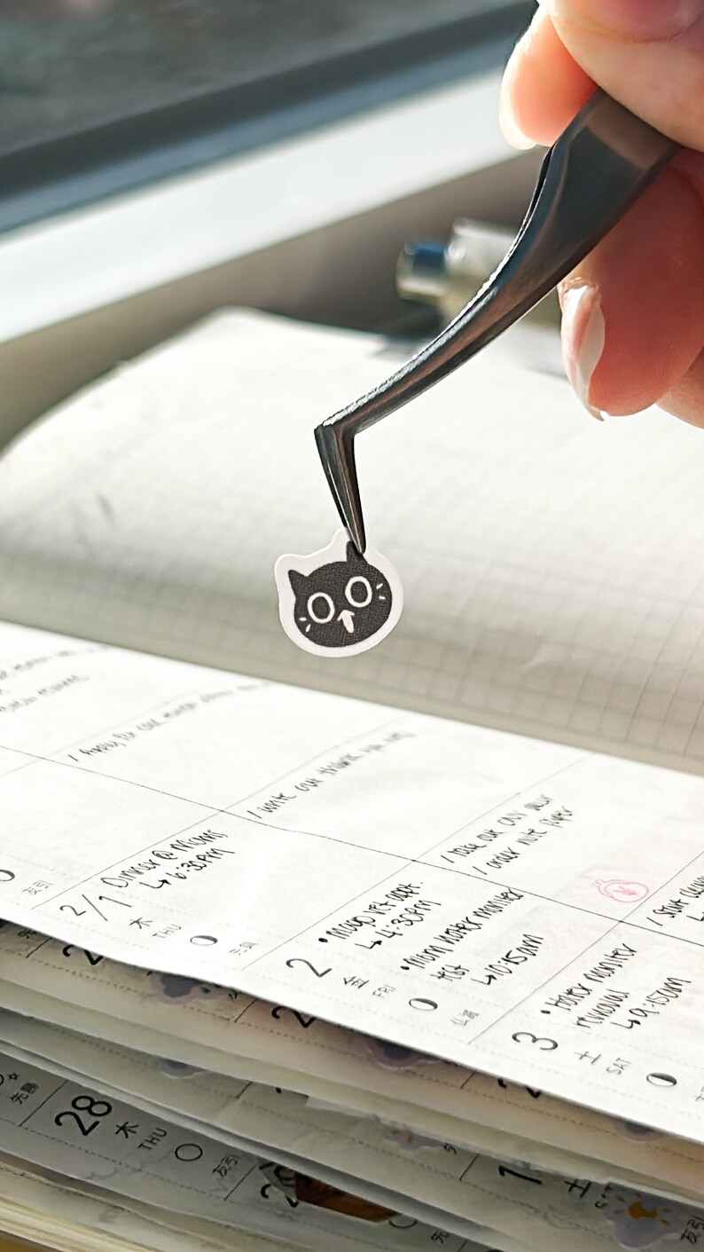 May include: A black and white cat sticker with large eyes being held by tweezers over a calendar.