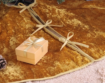 Puede incluir: Una falda de árbol de Navidad de terciopelo dorado con cierre de lazo a juego. Una pequeña caja de regalo con una cinta dorada se encuentra sobre la falda. Se ve una piña en la esquina inferior izquierda.