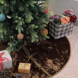 Puede incluir: Un &aacute;rbol de Navidad decorado con adornos, una falda de &aacute;rbol de terciopelo marr&oacute;n y regalos envueltos. La falda del &aacute;rbol es redonda con un ribete dorado y lazos de cinta. Una bolsa de regalo a cuadros con flores est&aacute; cerca.
