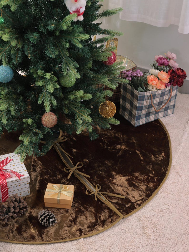 Puede incluir: Un &aacute;rbol de Navidad decorado con adornos, una falda de &aacute;rbol de terciopelo marr&oacute;n y regalos envueltos. La falda del &aacute;rbol es redonda con un ribete dorado y lazos de cinta. Una bolsa de regalo a cuadros con flores est&aacute; cerca.