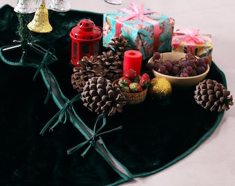Puede incluir: Una falda para árbol de Navidad de terciopelo verde oscuro con un diseño circular, una abertura y lazos de tela a juego. La falda está decorada con piñas, un farol rojo, una vela roja, un cuenco con uvas, fresas y regalos envueltos.