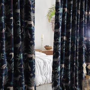 Puede incluir: Cortinas azul marino con un estampado de jungla. La tela presenta ilustraciones de leopardos, cebras, flamencos y palmeras en tonos verdes, blancos y beige. Las cortinas cuelgan frente a una pared blanca y una cama.