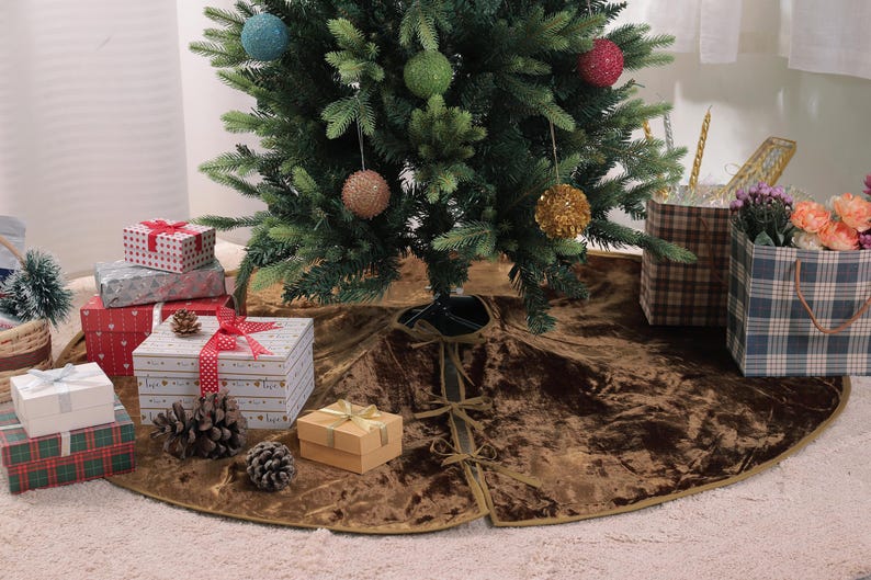 Puede incluir: Un &aacute;rbol de Navidad decorado con adornos coloridos se alza sobre una falda de &aacute;rbol de terciopelo marr&oacute;n. Regalos envueltos en varios tama&ntilde;os y patrones, junto con pi&ntilde;as, est&aacute;n dispuestos alrededor de la base, creando una escena festiva.