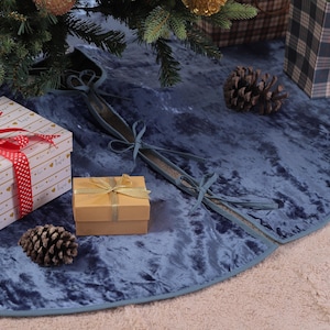 Op de afbeelding: Een blauwe fluwelen kerstboomrok met een lichtblauwe rand. De scène bevat geschenkdozen, dennenappels en een rood lint. Een kleine geschenkdoos is versierd met een gouden lint. De sfeer is feestelijk.