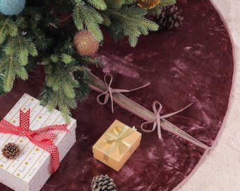 May include: A Christmas tree skirt in a deep burgundy velvet texture, with a pink trim. The skirt surrounds a decorated Christmas tree with ornaments and gift boxes wrapped in festive paper and ribbons. Pinecones add a natural touch.