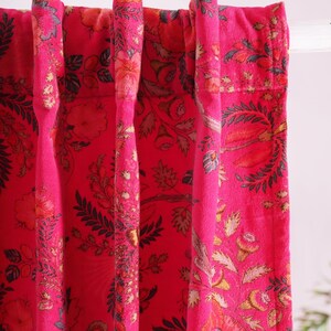 K&ouml;nnte beinhalten: Nahaufnahme von leuchtend pinkfarbenen floralen Vorh&auml;ngen. Die Vorh&auml;nge weisen ein detailliertes Muster aus Blumen und Bl&auml;ttern in Orange-, Gelb- und Gr&uuml;nt&ouml;nen auf. Der Stoff ist ein weiches, strukturiertes Material, das dem Design Tiefe verleiht. Die Vorh&auml;nge h&auml;ngen an einer wei&szlig;en Stange.