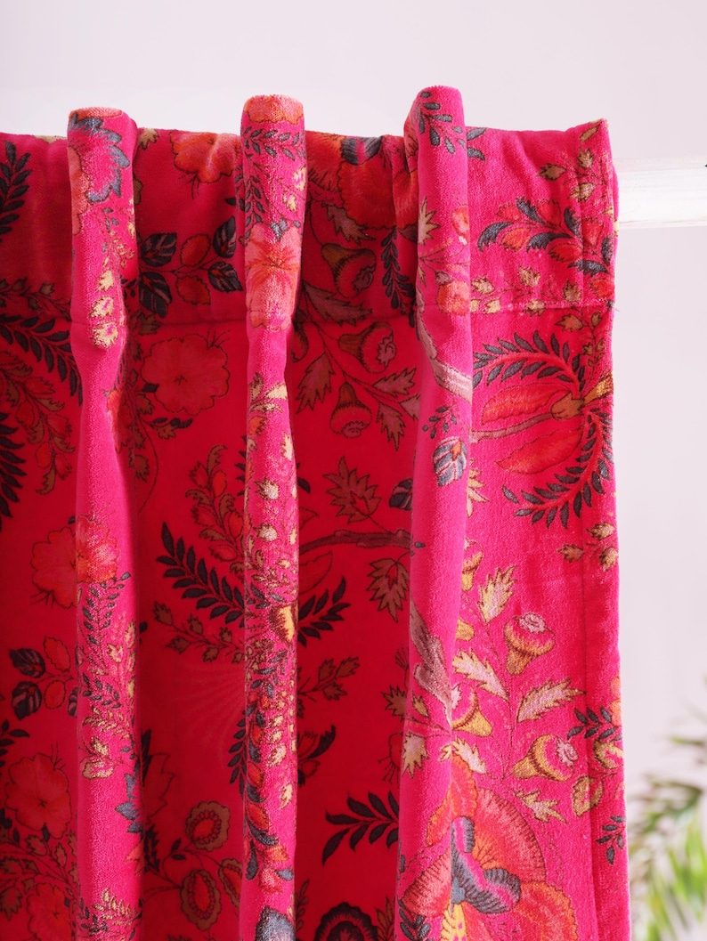 K&ouml;nnte beinhalten: Nahaufnahme von leuchtend pinkfarbenen floralen Vorh&auml;ngen. Die Vorh&auml;nge weisen ein detailliertes Muster aus Blumen und Bl&auml;ttern in Orange-, Gelb- und Gr&uuml;nt&ouml;nen auf. Der Stoff ist ein weiches, strukturiertes Material, das dem Design Tiefe verleiht. Die Vorh&auml;nge h&auml;ngen an einer wei&szlig;en Stange.