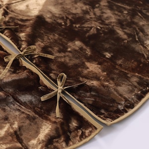 Puede incluir: Una falda de &aacute;rbol de Navidad de terciopelo marr&oacute;n con ribete dorado. La falda tiene una abertura dividida con lazos de cinta beige. El terciopelo tiene un aspecto suave y texturizado, y el dise&ntilde;o general es circular.