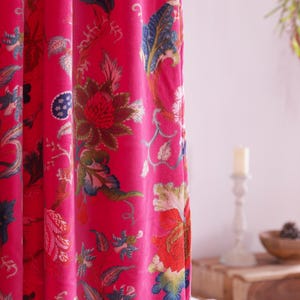K&ouml;nnte beinhalten: Leuchtend pinkfarbener Vorhang mit gro&szlig;em Blumenmuster. Der Stoff zeigt ein botanisches Design mit roten, blauen, gr&uuml;nen und cremefarbenen Blumen und Bl&auml;ttern. Der Vorhang besteht aus einem weichen, flie&szlig;enden Material und ist teilweise zugezogen.