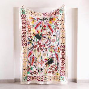 May include: A rectangular textile wall hanging featuring intricate, multi-colored floral and bird embroidery on a cream-colored background. The design includes a decorative border with repeating patterns in red, yellow, and green thread.
